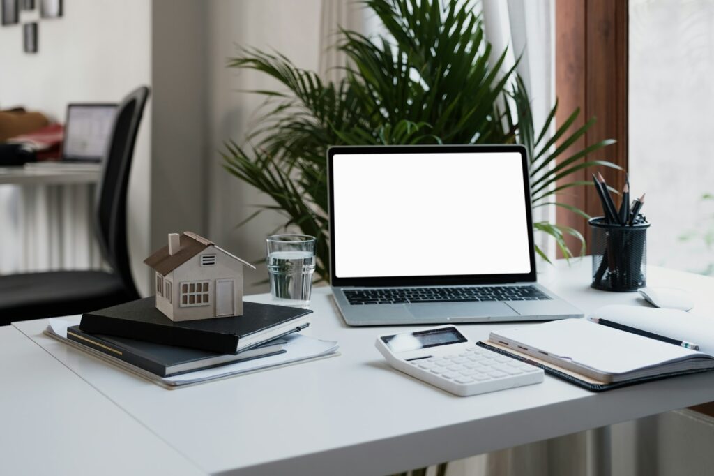 Laptop computer with blank white screen on modern desk, real estate with house model, notepad and